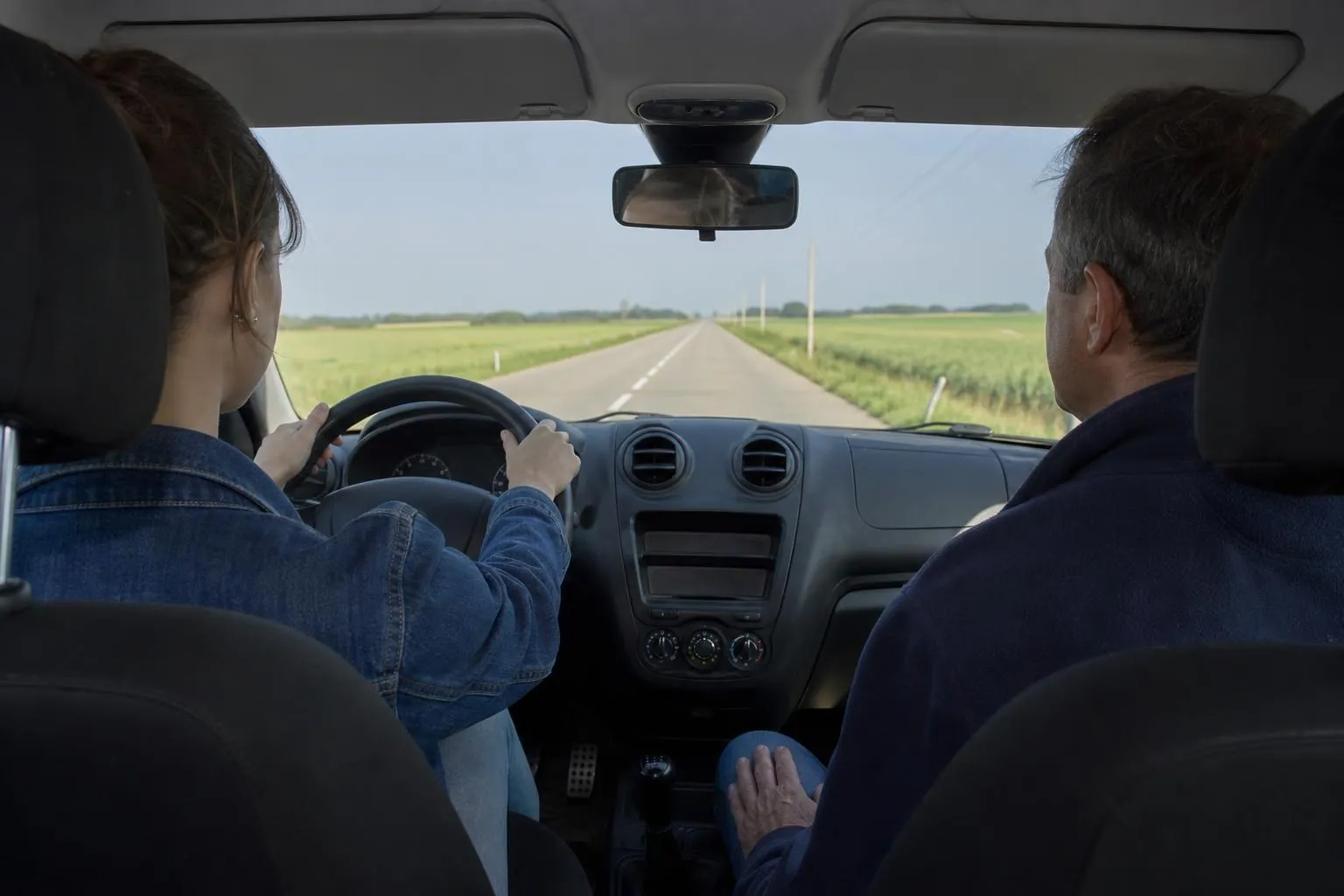 Vue depuis la banquette arriere d'une voiture ecole sur une route departementale bordee de champs, eleve au volant et enseignant a droite.