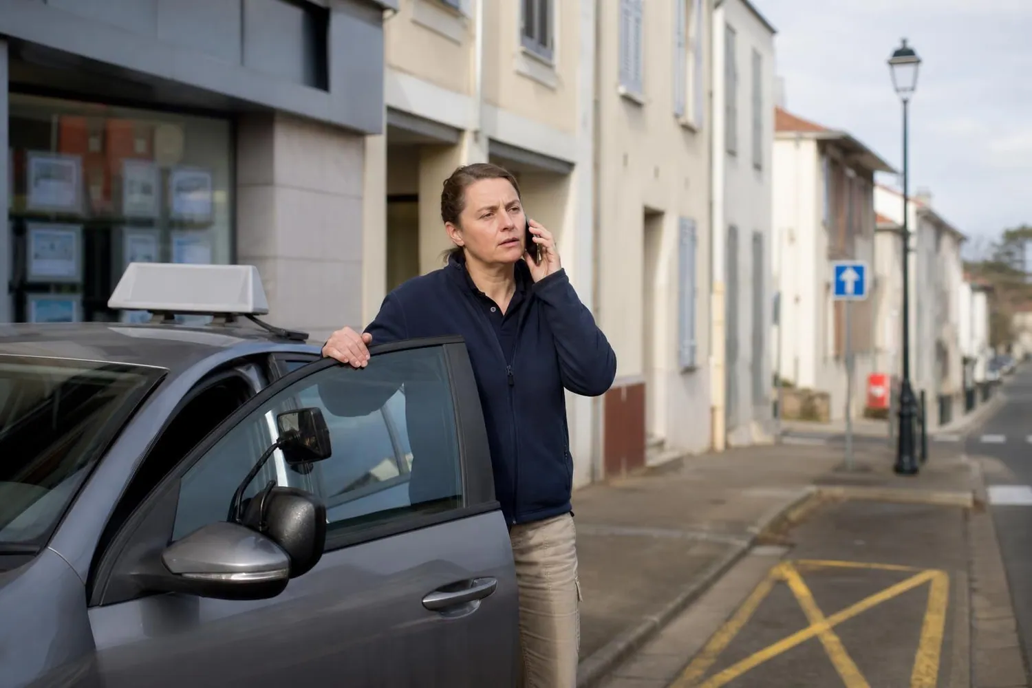 Gerante d&rsquo;auto ecole au telephone devant sa voiture ecole stationnee devant l&rsquo;agence d&rsquo;un bourg de campagne