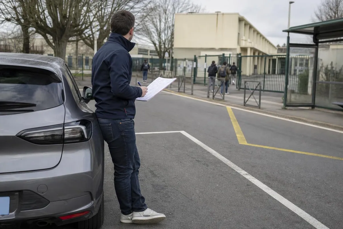 Enseignant de la conduite adosse a sa voiture ecole devant l&rsquo;entree d&rsquo;un lycee, feuille de planning a la main